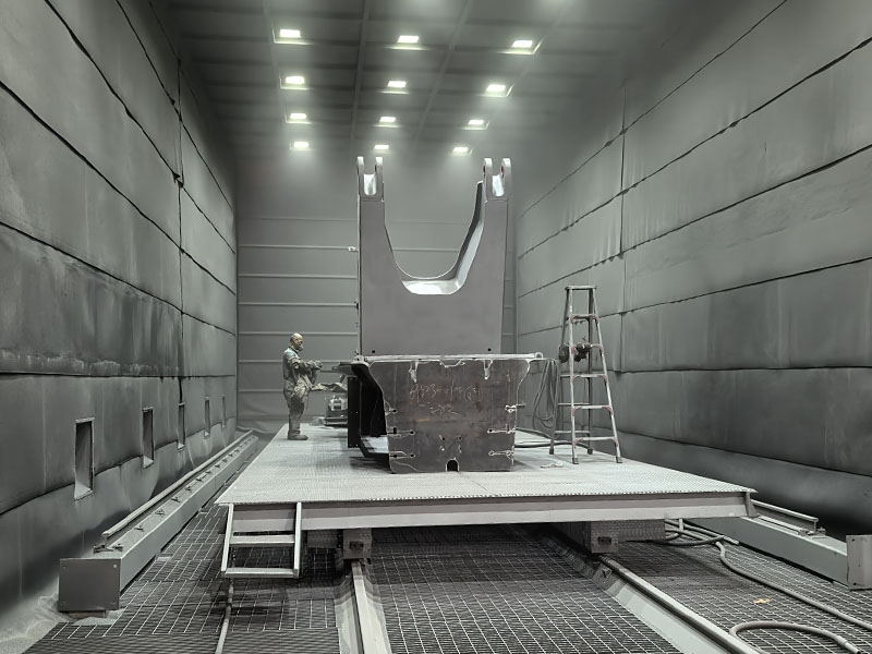 Manual sandblasting chamber operation on heavy steel H-beams using metallic grit abrasive media in a shipyard shot blasting booth.