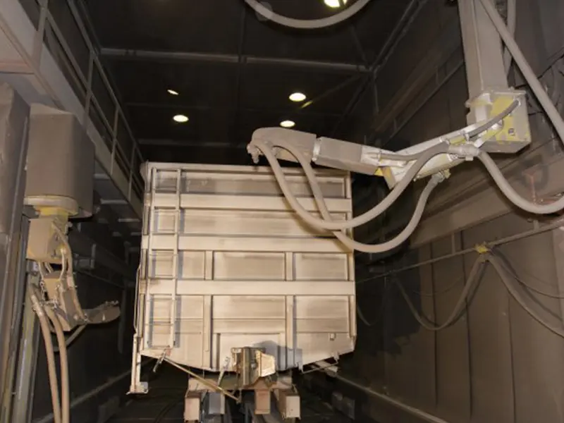 Overhead gantry-mounted robotic blasting system performing automated surface preparation on a heavy railway wagon chassis inside the chamber.