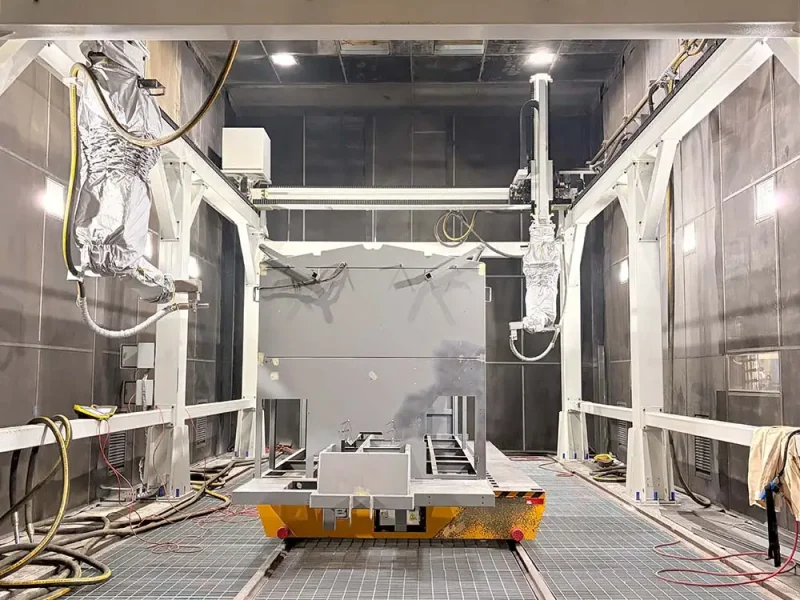 Dual gantry-mounted blasting robots collaborating inside the chamber with a heavy-duty yellow transfer cart in the center.
